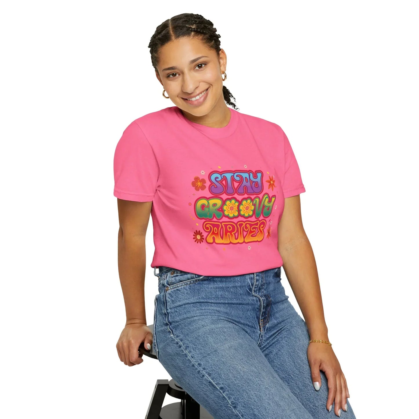 Smiling woman in braided hair wearing pink T-shirt with colorful Stay Groovy Aries graphic, sitting on stool