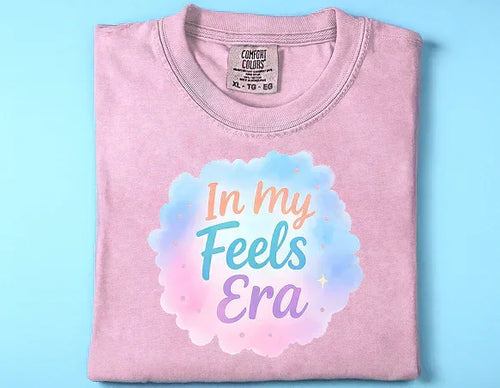 Folded pink Comfort Colors t-shirt with pastel cloud graphic and text In my Feels Era on blue background